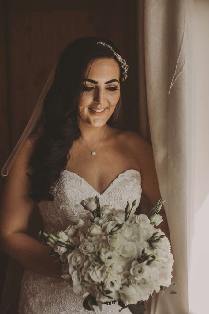 portrait of bride in wedding dress, looking at bridal bouquet of flowers lit with natural window light