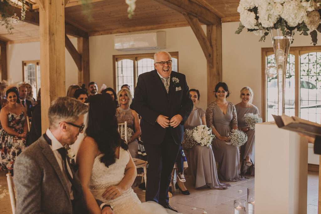 the bride sits with the groom during wedding ceremony as grooms father looks on at Colshaw Hall